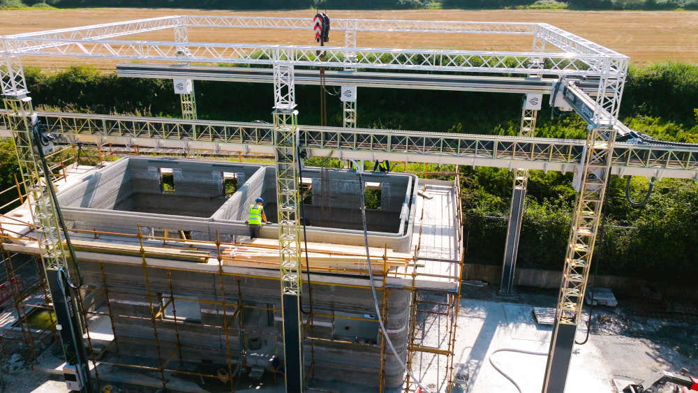 Aerial view of a 3D construction printer in operation, building the concrete structure of a two-story house with workers on scaffolding around the site.